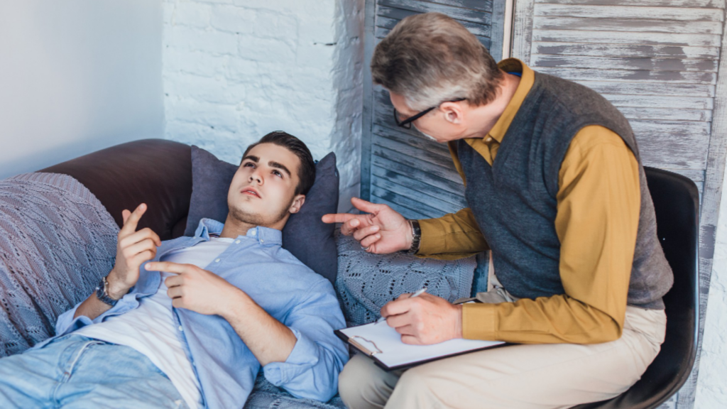 A young gay man lying on a couch during a therapy session, while an older male therapist sits nearby, taking notes and gesturing as they engage in conversation